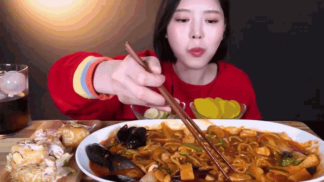a woman in a red sweater is eating a plate of food with chopsticks .