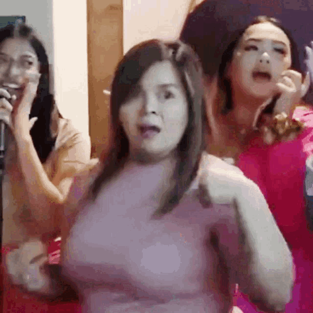 a woman in a pink dress is singing into a microphone while two other women are talking on their cell phones .