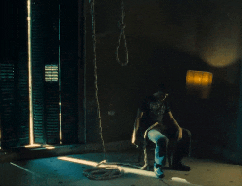 a man is sitting in a dark room with a noose hanging from the wall