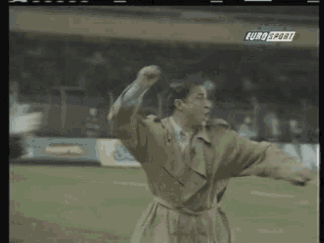 a man in a trench coat stands on a soccer field with his fist in the air in front of a euro sport sign