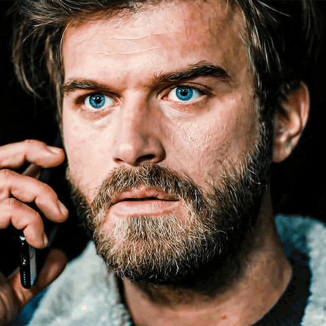 a man with a beard and blue eyes talking on a cell phone