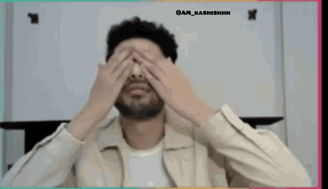 a man with a beard is covering his eyes with his hands while wearing a jacket .