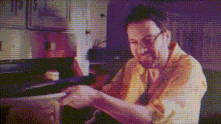 a man in a yellow shirt is sitting in a kitchen looking at something