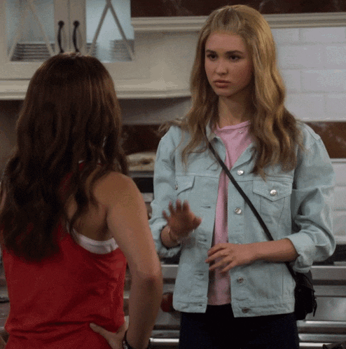a woman in a blue denim jacket talks to another woman in a red tank top