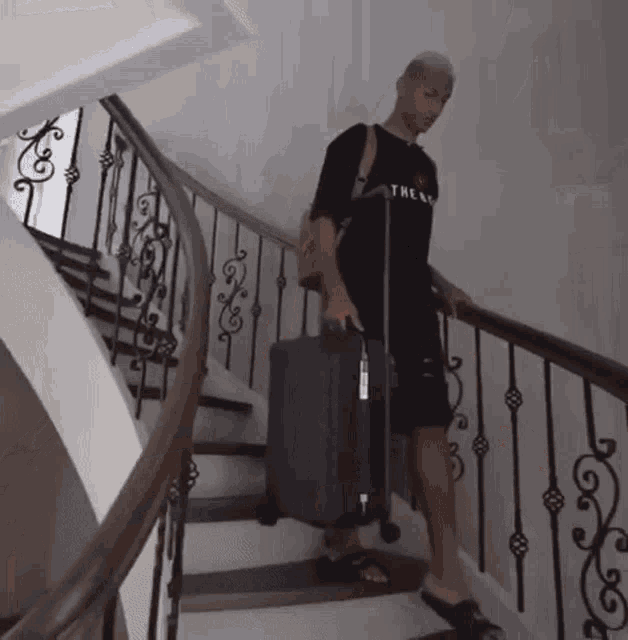 a man wearing a black shirt with the word the on it is carrying a suitcase up a set of stairs .