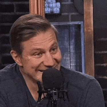 a man laughs in front of a microphone with a brick wall behind him