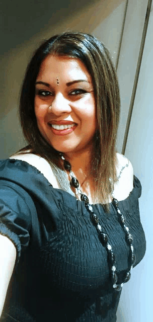 a woman wearing a black off the shoulder top and a black necklace smiles for the camera