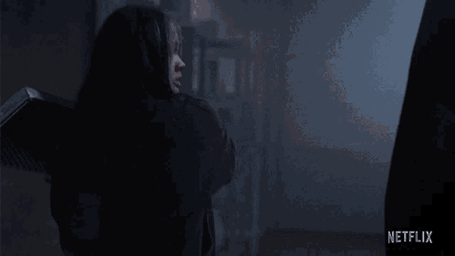 a woman in a black hoodie is standing in a dark room with a netflix logo in the corner .