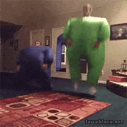 two people in green jumpsuits are jumping in a living room .