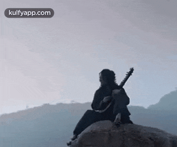 a man is sitting on a rock playing a guitar in the mountains .