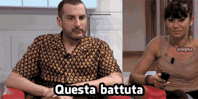 a man is sitting next to a woman who is holding a cell phone and says questa battuta