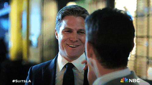 a man in a suit and tie smiles while talking to another man with #suitsla written on the bottom