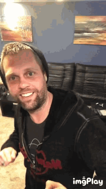 a man with a beard is smiling in front of a couch and a painting on the wall behind him