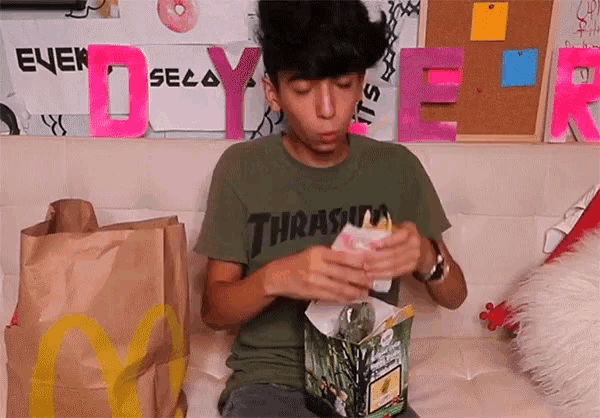 a man wearing a thrasher shirt is sitting on a bed eating a sandwich