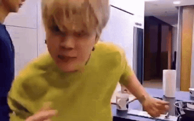 a man in a yellow shirt is standing in a kitchen next to a cup of coffee .