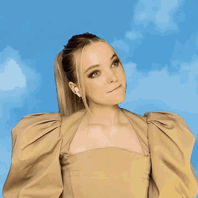 a woman wearing a tan dress with puffed sleeves is standing in front of a blue sky with clouds .