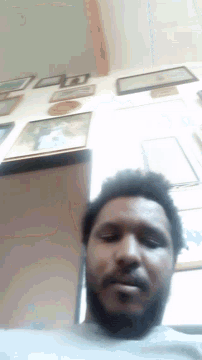 a man with a beard is looking at the camera in front of a wall with pictures on it
