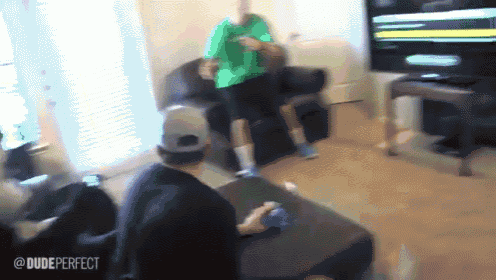 a man in a green shirt is playing a video game while another man sits on a couch