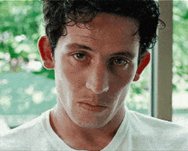 a man with curly hair is wearing a white t-shirt and looking at the camera with a serious look on his face .