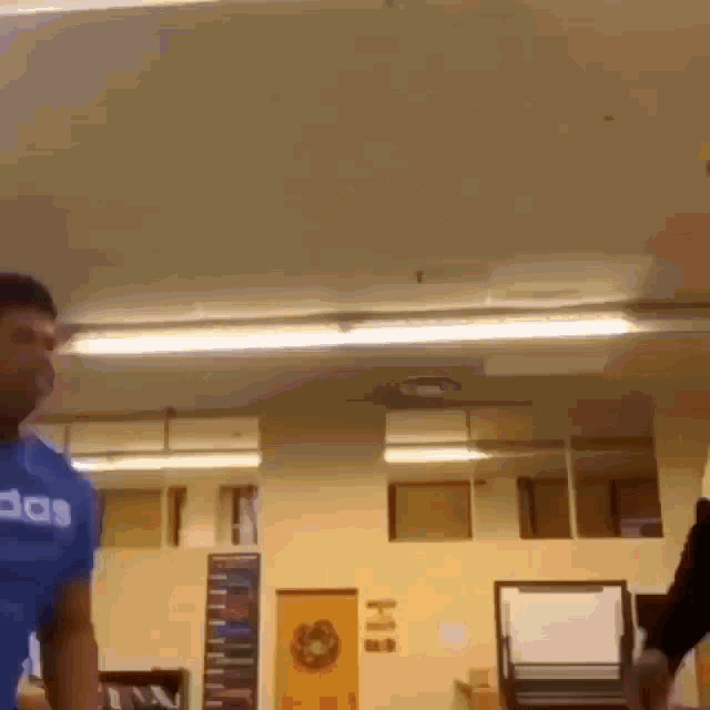 a man in a blue adidas shirt is standing in a room with a ceiling fan .