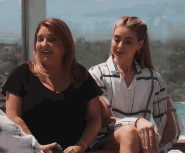 two women are sitting next to each other in front of a window and smiling