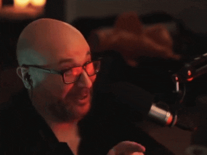 a bald man with glasses and a beard is pointing at the camera in front of a microphone .