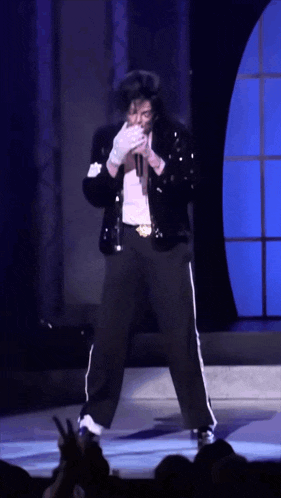 a man in a black jacket and white gloves is singing into a microphone on a stage