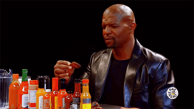 a man in a leather jacket is sitting at a table with many bottles of hot sauce