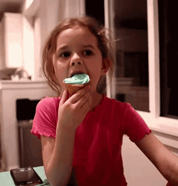 a little girl in a pink shirt is eating a cupcake with a green frosting
