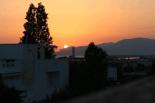 a sunset with a building in the foreground and the sun shining through the trees