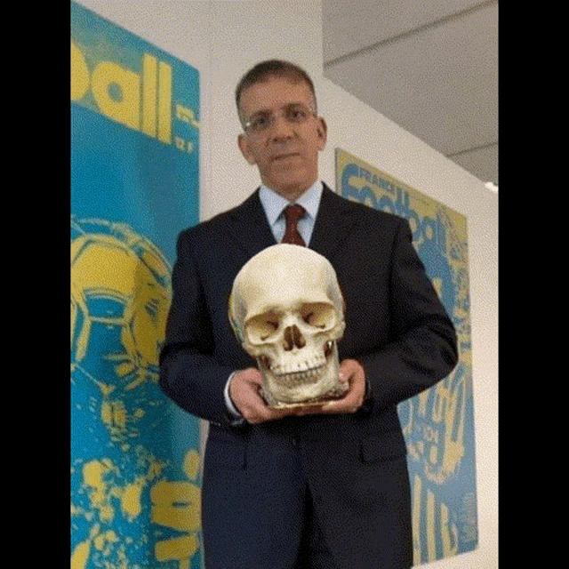 a man in a suit and tie is holding a skull in front of a wall with a poster that says ball