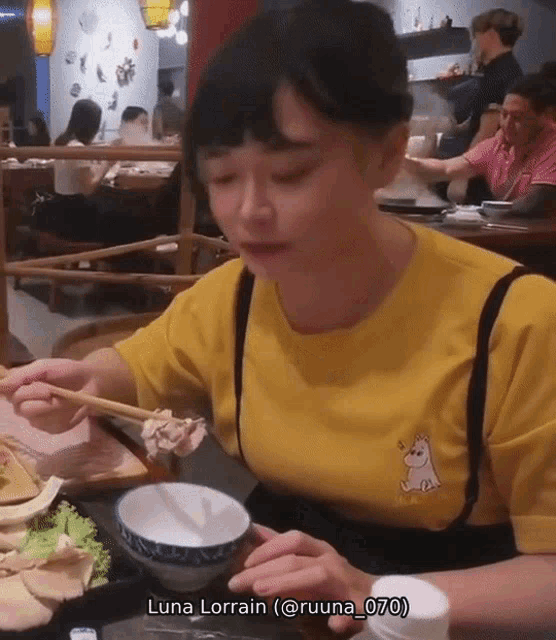 a woman wearing a yellow shirt with a unicorn on it is eating food with chopsticks ..