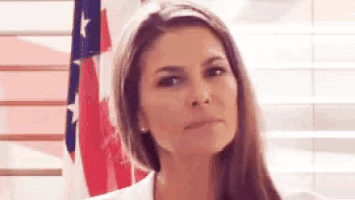 a woman in a white coat is standing in front of an american flag and making a funny face .