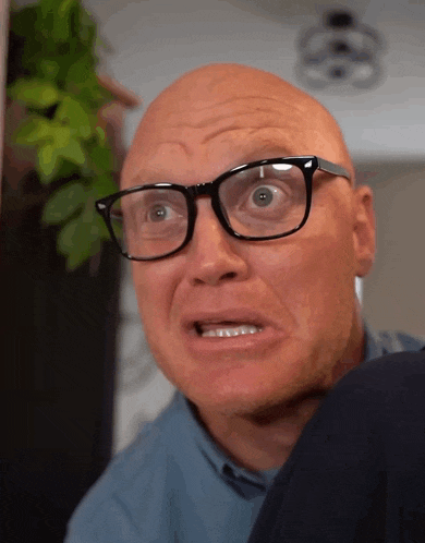 a bald man wearing glasses and a blue shirt makes a funny face
