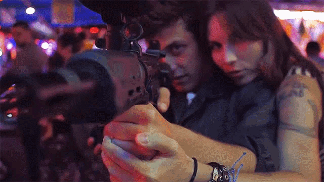 a woman with a tattoo on her arm looks at a man aiming a gun