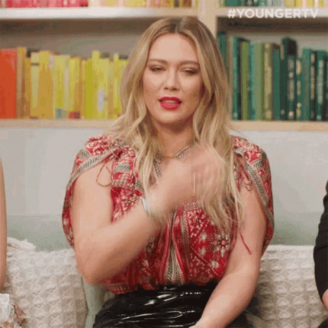 a woman in a red top and black skirt is sitting on a couch in front of a book shelf .