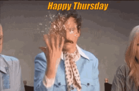 a man with a mustache is sitting in front of a group of people with a happy thursday greeting .