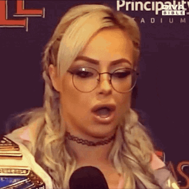 a woman wearing glasses and a choker is talking into a microphone in front of a sign that says principality bible
