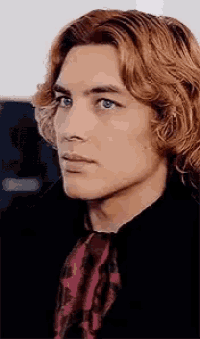 a young man with long red hair and blue eyes is wearing a black jacket and a red scarf .