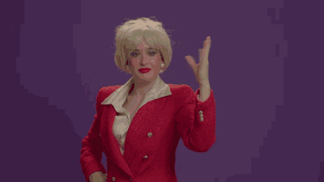 a woman in a red jacket and blonde wig waves her hand