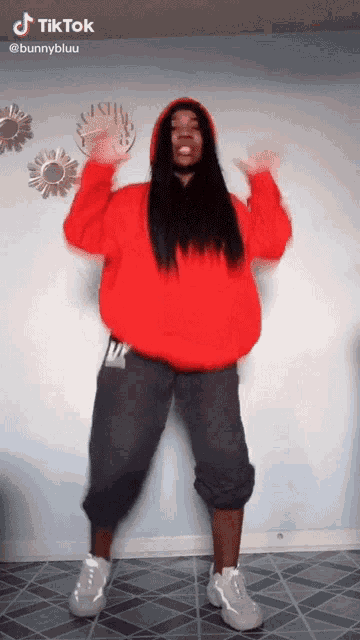 a woman in a red hoodie is dancing in front of a wall with a clock on it