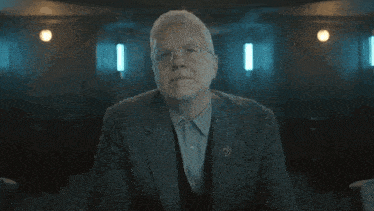 an older man in a suit and tie is sitting with his arms crossed in a dark room .