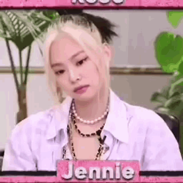 a woman wearing a white shirt and a pearl necklace is sitting in front of a pink sign that says jennie .
