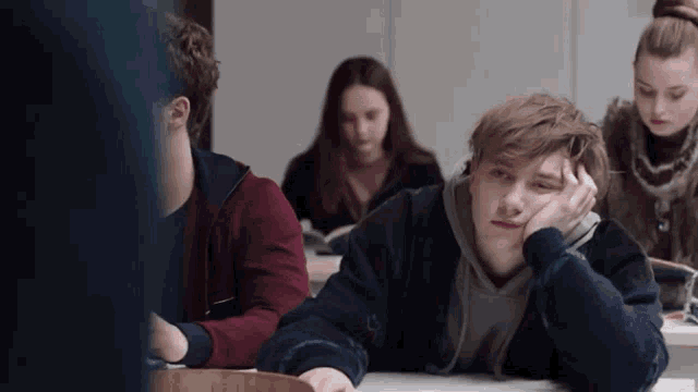 a group of young people are sitting in a classroom with one boy laying down with his head on his hand .