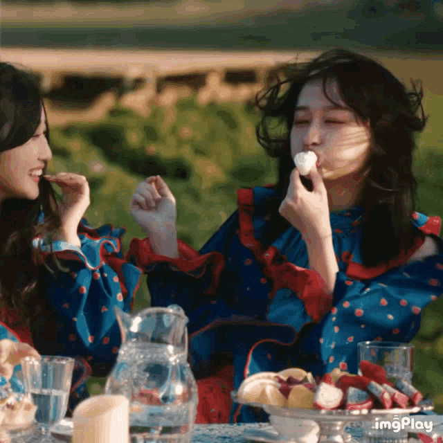 two women are sitting at a table with a pitcher of water and a plate of food and one of them is eating a piece of food