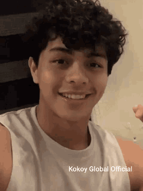 a young man with curly hair is wearing a white tank top that says kokoy global official on the bottom