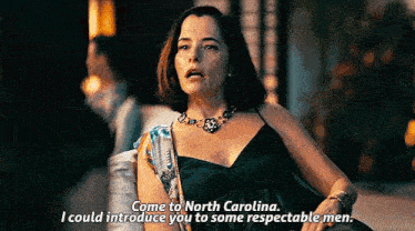 a woman in a black dress and necklace is talking to a man in a dark room .