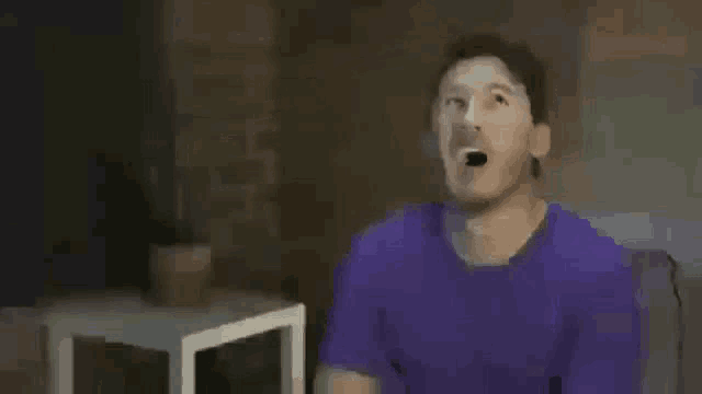 a man in a purple shirt is singing into a microphone while sitting on a couch .