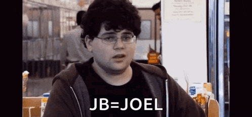 a man wearing glasses and a hoodie is sitting at a table with the words jb = joel written on it .