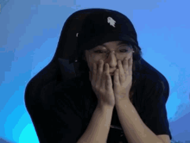 a woman wearing a black hat and glasses is sitting in a chair with her hands on her face .